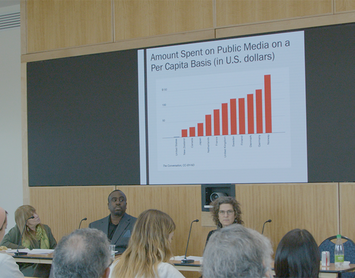 People seated at a table facing an audience, in front of a slide with the headline &quot;Amount spent on public media on a per capita basis (in US dollars)&quot;