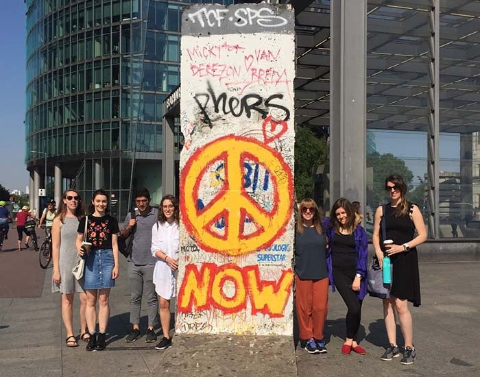 Seven people standing next to a chunk of the Berlin Wall with a graffiti peace sign and the word &quot;now&quot;