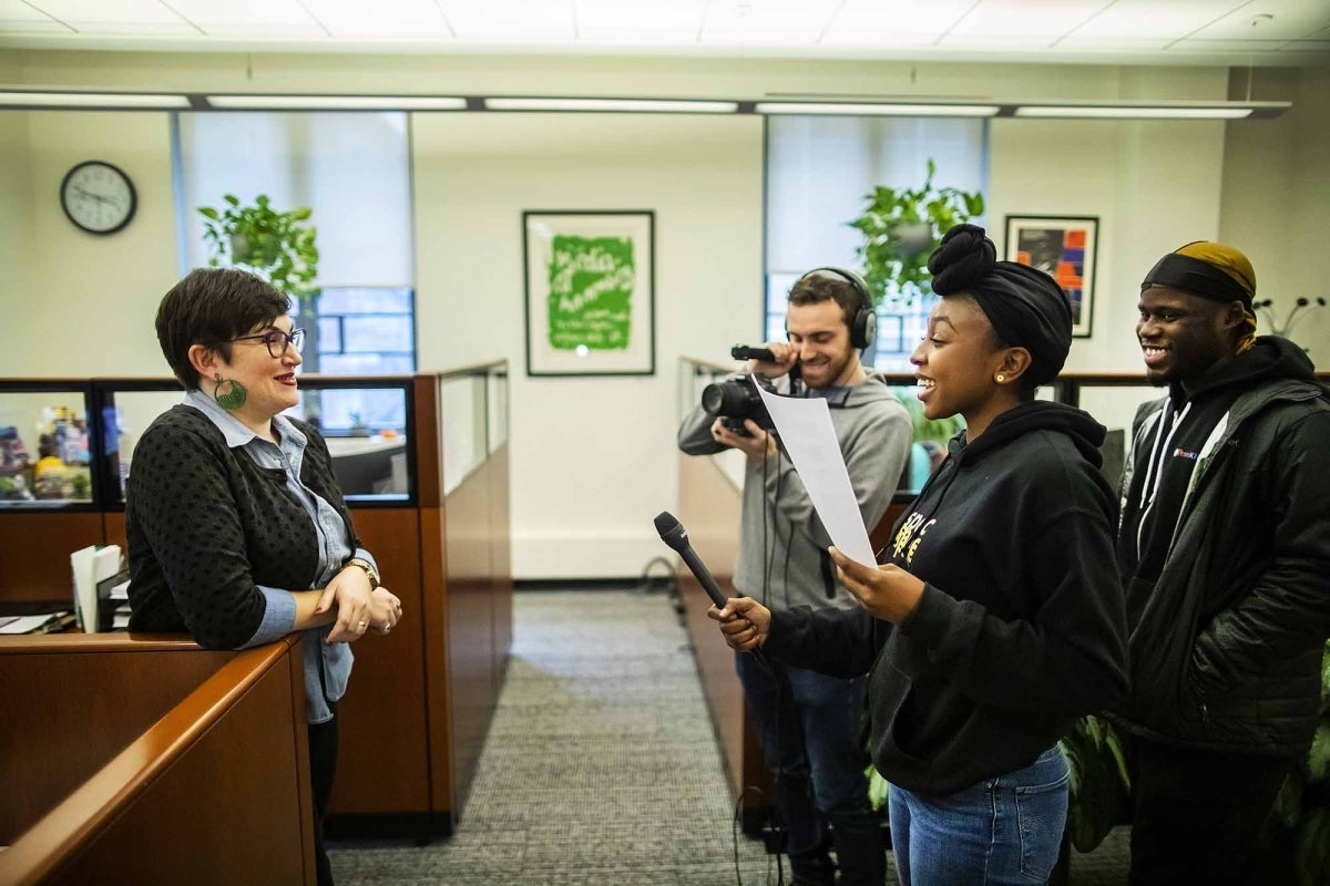 Student holds microphone while interviewing woman. Another student holds a camera while a third watches.