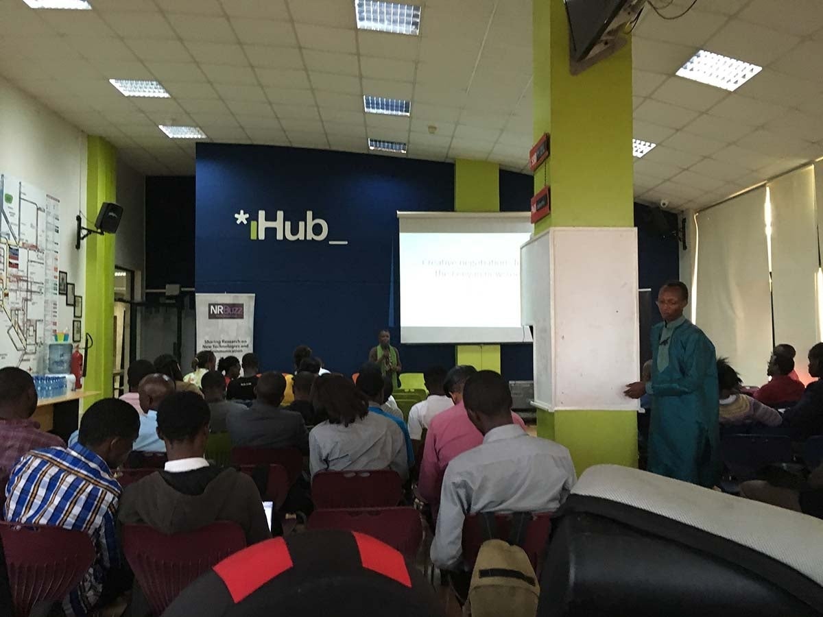 Room with light green, white and dark blue walls filled with many men seated towards the front where someone, with the help of a powerpoint, is giving a presentation. The front wall, close to the ceiling, has an iHub sign, with the 'I' in green and 'hub' in white.