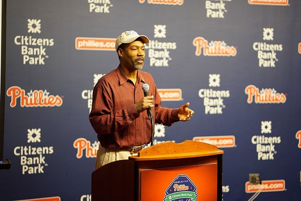 Glanville lecturing of the semester in the Phillies media room. He is standing behind a low, wooden podium, and infront of a blue backdrop with Citizens Bank Park and the Phillies' logos in an alternating tile pattern.