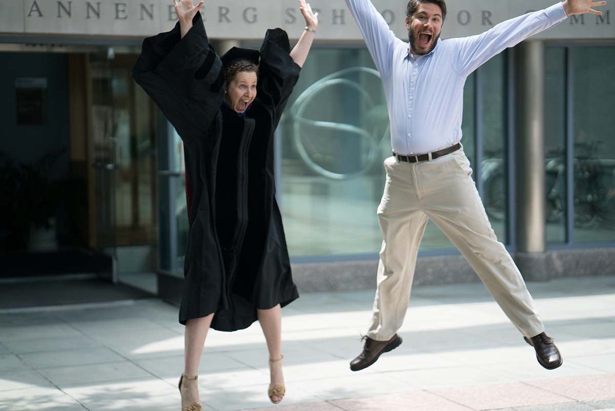 Elissa Kranzler, in her graduation wear, and her partner jumping for joy in front of the Annenberg School for Communication.