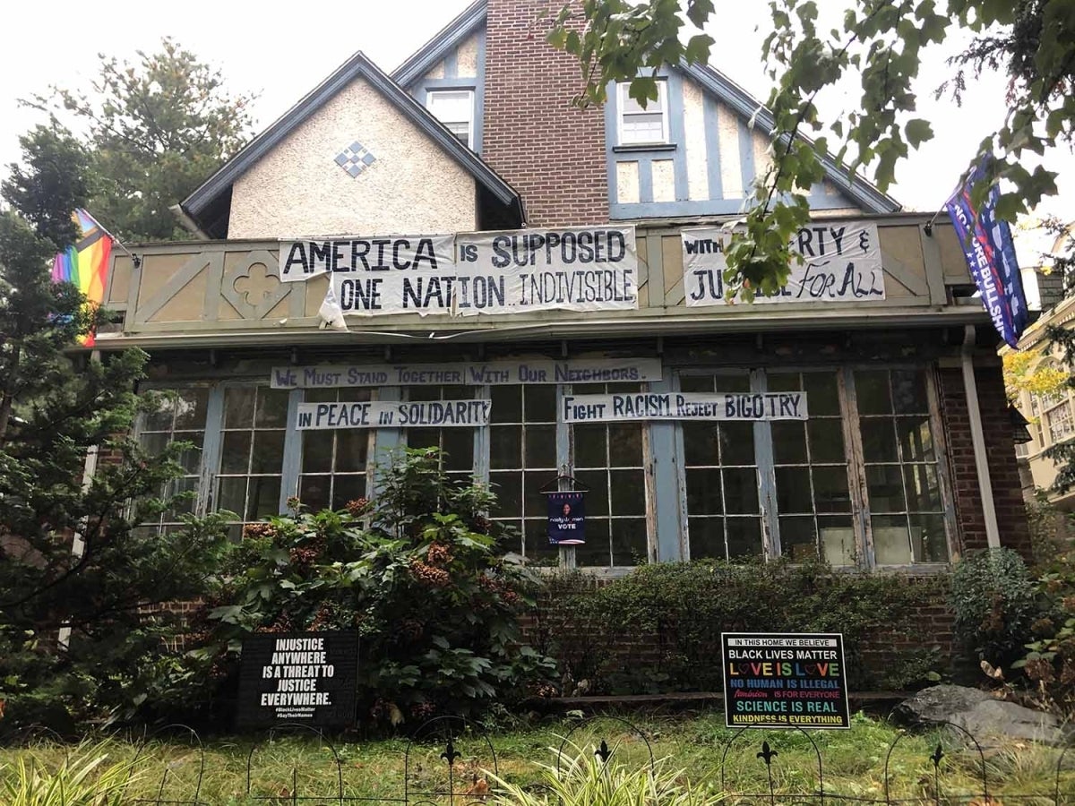 House with many signs and flags. Some are handmade and some are not. One flag says “No more Bullshit” under something else that is not legible in the picture. Another has the Philadelphia LGBTQ pride flag. One of the large handmade banners reads “America is supposed” then “One nation indivisible” with one piece folded down (presumably saying “be”) another banner reads “with liberty & justice for all”. A banner below it says “We must stand together with our neighbors. In peace in solidarity. Fight racism, re