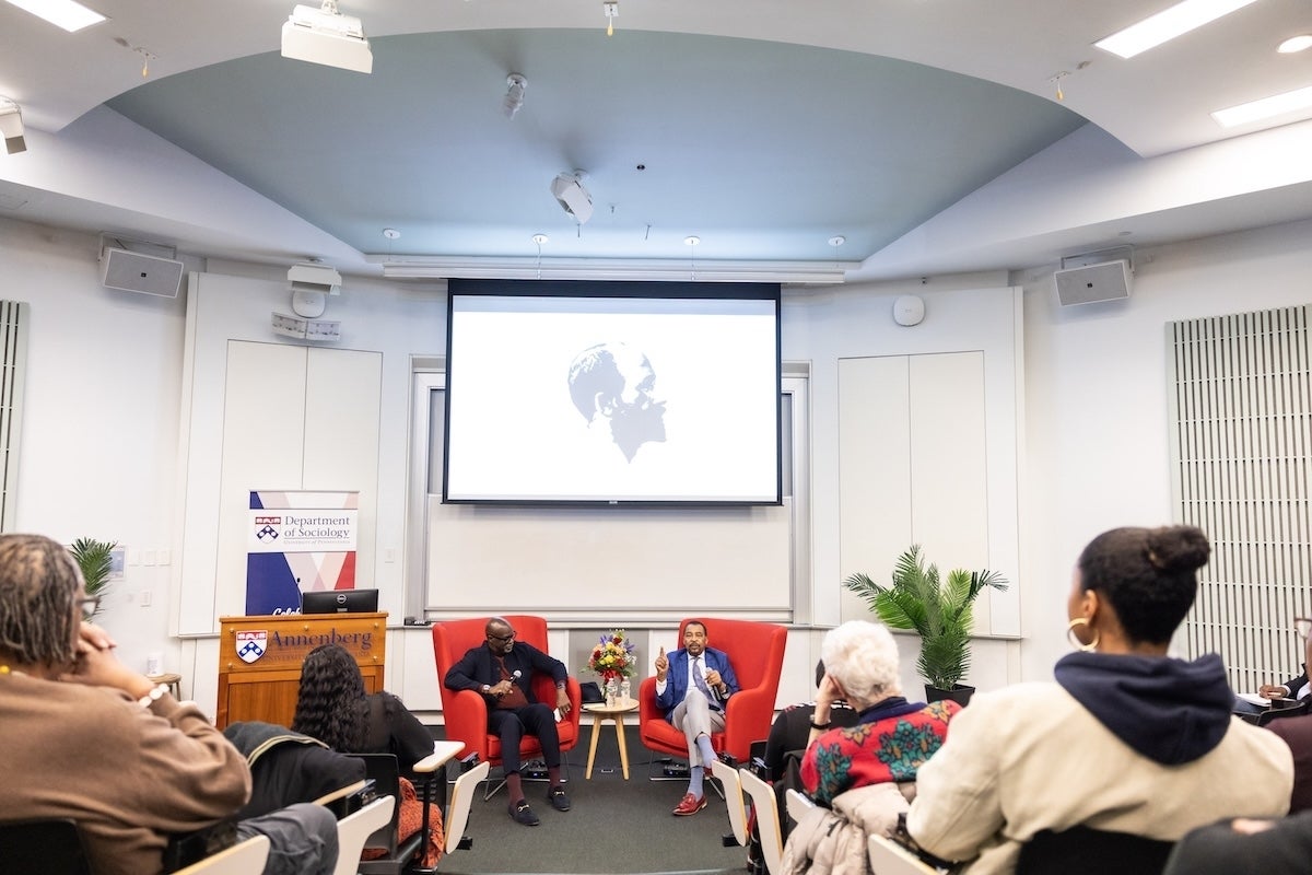 Tukufu Zuberi and Aldon Morris sit in chairs in the front of an audience in an auditorium and talk to one another