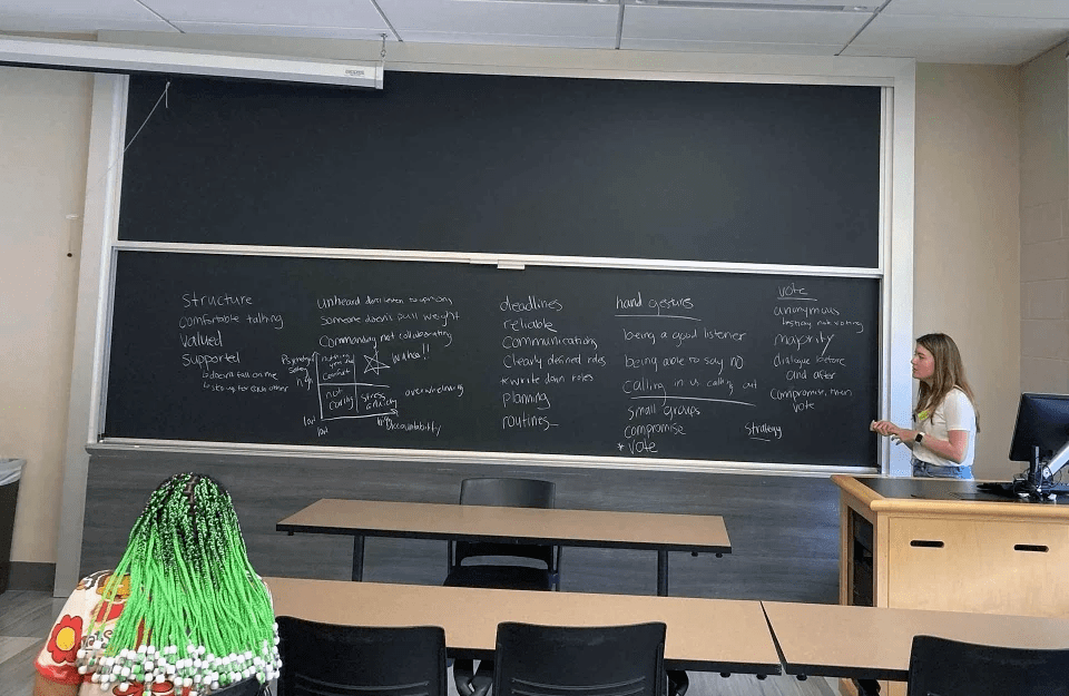 Teacher standing by a chalkboard filled with words. In the classroom, we see the back of a student's head