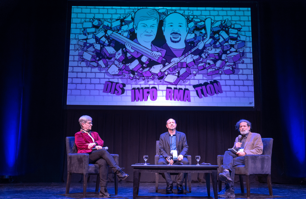  Jamieson, Mann, and Weiss sit on stage during opening plenary discussion, with slide on screen behind them featuring a cartoon of Jamieson holding a pencil and Mann holding a hockey stick breaking through a brick wall labeled "DIS-INFO-RMA-TION."