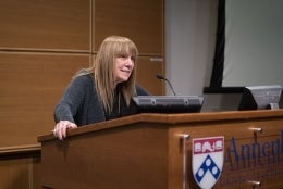 Barbie Zelizer stands behind a large desk with a microphone. She is delivering a speech. The desk is made of wood and in front of her is a black monitor. The front of the desk has the University of Pennsylvania shield with the word Annenberg beside it. 