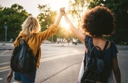 Two young women from the back, holding hands with arms raised as they walk in the street at sunset