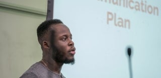 Student speaking with a screen behind him reading "Communication Plan"