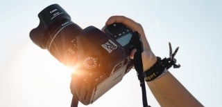 A right hand holds up, at an angle, a professional camera with an attached lens up to the sky. Photo by Christian Wiediger on Unsplash.