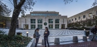 Locust Walk side panorama of the Annenberg School building with two students speaking to one another in the foreground. It appears to be dusk.