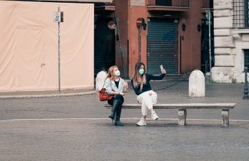 Two women sit on bench and take a selfie. Both are wearing face masks to protect against Covid-19.