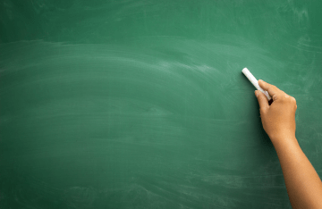 Hand holding chalk next to a green, empty chalkboard