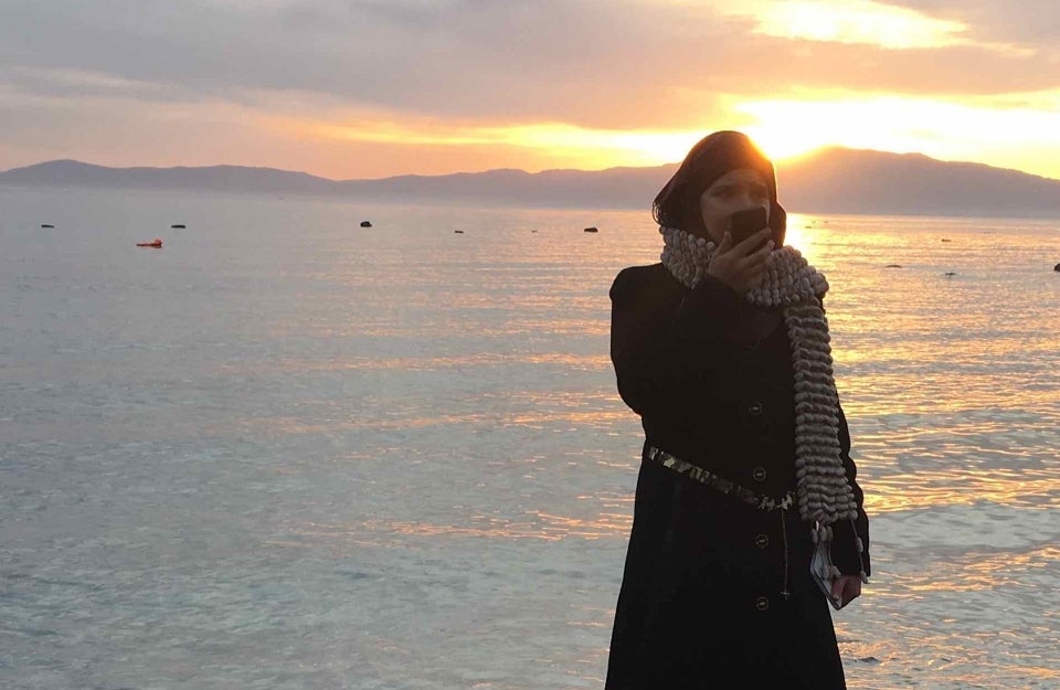 This photo is taken at sunset or dawn, with the sun just above the hills that are to the right. Infront of these hills is the ocean that reflects the sunlight. In the foreground is. woman standing on the beach wearing a long dark coat, a scarf and a head covering. She is speaking on the phone.