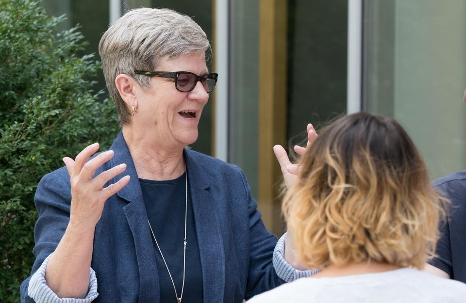 Kathleen Hall Jamieson outside speaking to people. Her hands are up as she speaks. She is wearing black/transition glasses, a black shirt, a navy blue blazer and a long silver necklace.