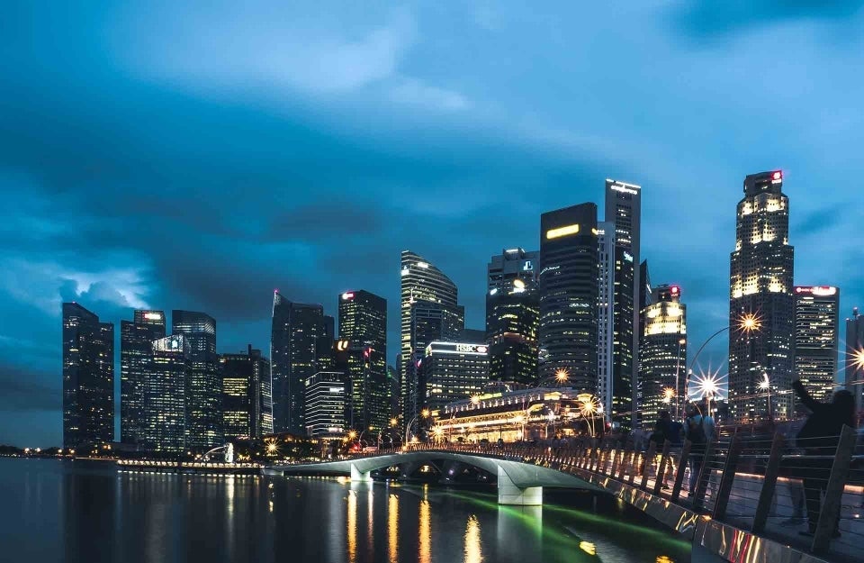 Taken at twilight with a cloudy sky, this is a city with numerous tall buildings, each illuminated by their respective artificial driving. The buildings are in the background and before them is a bridge above a body of water. There are people on the bridge. photo credit Shawn Ang / Unsplash