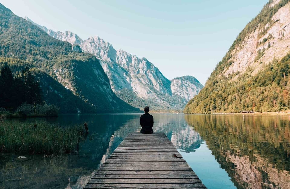 Boardwalk leading into a water body. At the edge of the walk is a person sitting down. The water body is surrounded by green mountains. photo credit Simon Migaj / Unsplash