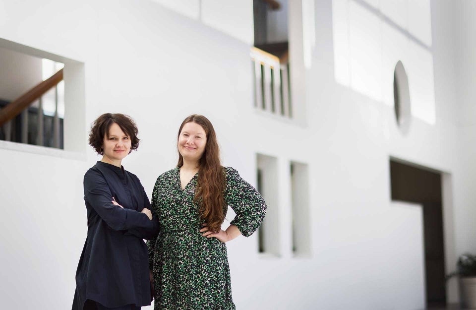 Two women posing together in front of a white wall with various architectural cutouts