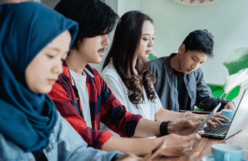 Four people sit next to one another at a cafe table looking down at their digital devices