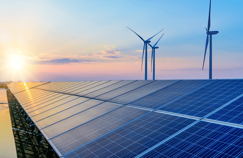 a landscape with a sunset shows rooftop solar panels in the foreground and wind turbines in the background