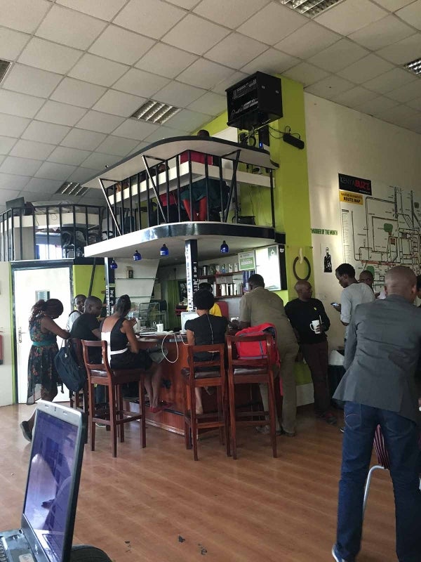 Cafe with people sitting on brown chairs using computers or speaking with one another. Two walls are painted light green and one white.