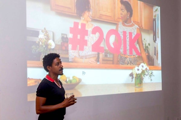 Aymar Jean Christian at the premiere of "Two Queens in a Kitchen". He stands in front of a projected image that has a picture of two people standing in a kitchen as they converse. In the center of the image in big, pink text is '#201K'.