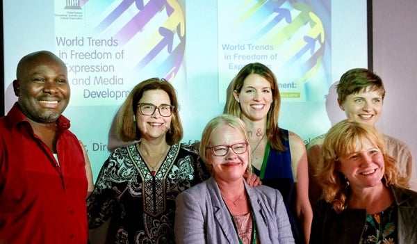 Jennifer Henrichsen poses with fellow panelists in front of the projector screen. They are two rows of people. The projector is still on. 