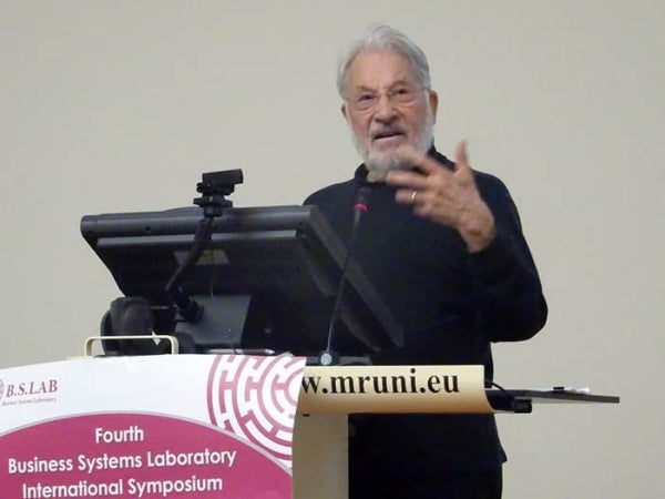 Klaus Krippendorff speaking behind a podium. There are a black monitor and a black mic on the podium.