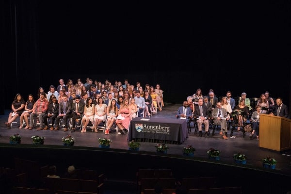 Graduates and faculty members seated in neat rows on a darkened theater stage, ready for graduation to begin