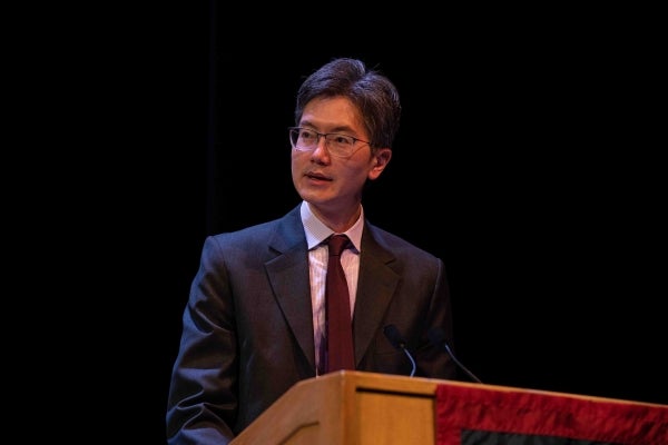 Andy Tan wearing a suit and speaking at a podium with a black background