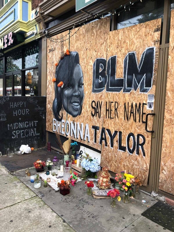 A memorial for Breonna Taylor. It is painted on a boarded up building and reads “BLM Say Her Name Breonna Taylor” in black and white and has a portrait of her. Her hair has real flowers attached to it. There are flowers and candles below the memorial forming an altar. The sidewalk has Black is Beautiful painted on it with an abstract face next to it (same as featured on the dumpster). 