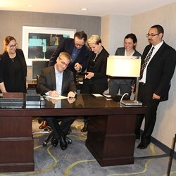 Professor Kathleen Hall Jamieson, her doctoral students, and alumni Paul Waldman in a room together. Waldman is sitting behind a wooden desk focused writing something on a yellow paper as the others look at him intently.   