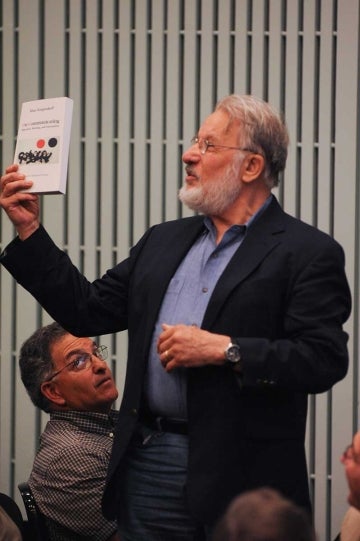 Man wearing a suit holds up a book with another man looking up at hime and wood slats in the background.